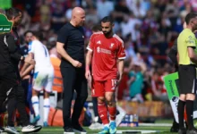 محمد صلاح واقفا على أرض الملعب بقميص ليفربول وهو يمسك ساقه متألماً، بينما يحيط به الجهاز الطبي لتقييم إصابة صلاح.